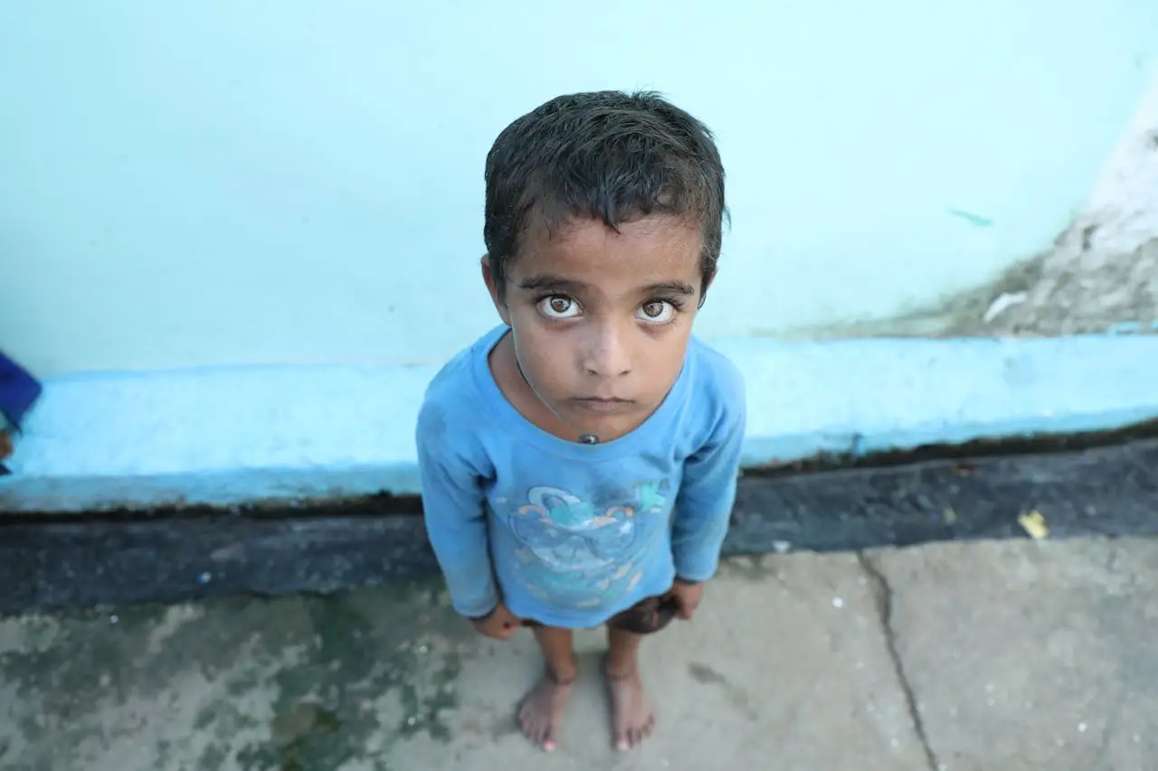 A young boy in Mungeli, India, gazes upwards on a concrete street, barefoot and wearing a blue shirt.